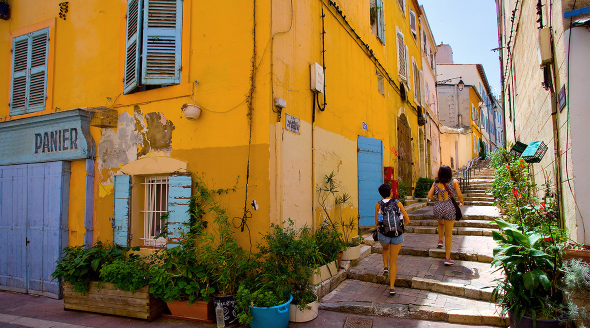 A bright yellow building with blue shutters and doors with a sloped alleyway on the right with two women in shorts heading up